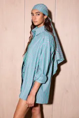 Camisa holgada de algodón con rayas verticales en tonos verde agua, rosa pálido y blanco. Presenta una abertura frontal que llega hasta la mitad del torso, ribeteada con un cordón ondulado de color verde brillante. Incluye botones oscuros y un pañuelo a juego en la cabeza.