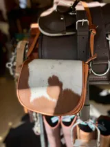 Bandolera de cuero con solapa de pelo vacuno en tono marrón y blanco, cuerpo de cuero marrón, correa larga regulable y detalles metálicos.