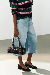 Bolso estilo bowling de cuero sintético acharolado color marrón, con diseño cilíndrico, bolsillos laterales con cierre y doble asa decorada con tachas metálicas doradas.