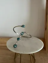 Brazalete abierto de estilo minimalista, confeccionado en metal plateado con textura martillada. Presenta un diseño espiralado con cuatro terminales decorativos circulares que contienen incrustaciones de piedras en tono turquesa con destellos brillantes.