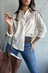 Camisa de color beige con textura rayada vertical, cuello clásico y mangas largas. Presenta un delicado bordado floral en tono sobre tono en la parte superior delantera.