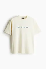 Remera de algodón color beige con cuello redondo y diseño holgado, con estampa frontal centrada con el texto "Enjoy the present" en azul.