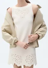 Vestido corto de tejido de punto fino color crema, con cuello barco y sin mangas. Presenta un diseño recto con detalles calados en forma de flores en el pecho y en el ruedo, el cual tiene un acabado festoneado.