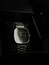 Reloj Casio con caja cuadrada de bordes redondeados, esfera negra minimalista con índices lineales, manecillas plateadas y ventana de fecha. Cuenta con malla de acero inoxidable de eslabones ajustables.