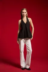 Blusa de algodón color beige con escote en V, cuello halter y cierre frontal con botones. Presenta un diseño ajustado en el torso con pinzas y pliegues en la parte inferior que generan una caída ligeramente acampanada.