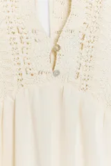 Vestido largo color crudo, sin mangas, con escote en V pronunciado, canesú superior de tejido de punto con botones frontales y lazo para atar en el cuello, y falda fluida con detalle de bordado en el bajo.
