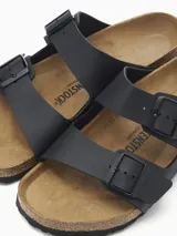 Sandalia de dos tiras ajustables con hebillas metálicas, color negro mate. Presenta la clásica plantilla anatómica de corcho y látex con forro de ante y suela de EVA.