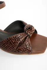 Sandalias de cuero marrón con taco cuadrado medio, tira en el tobillo con hebilla ajustable y diseño trenzado con nudo en la parte delantera.