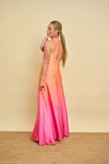 Vestido largo de satén con diseño degradado en tonos salmón y fucsia. Presenta cuello halter con detalle de nudo, falda evasé con tajo frontal pronunciado y espalda descubierta.