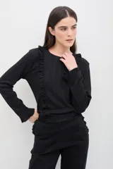 Remera de manga larga color negro, confeccionada en tejido suave con textura acanalada. Presenta volados verticales en el frente y terminaciones delicadas con volados en los puños.