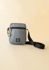 Bandolera gris con correa negra ajustable y logo de la marca Rusty en la parte frontal.