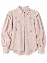 Camisa de color crudo con cuello camisero clásico, mangas abullonadas con puños estructurados terminados en ondas y delicados bordados florales en la parte frontal.