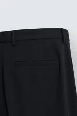 Pantalón negro de corte recto (regular fit) confeccionado en tejido compacto. Presenta pliegues frontales marcados, cierre frontal con botón y cremallera, y bolsillos de vivo en la parte trasera.