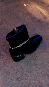 Bota corta de velvet azul marino con efecto abullonado en el tobillo y cierre lateral con cremallera. Tiene un tacón cuadrado y una cadena desmontable con el logo de la marca.