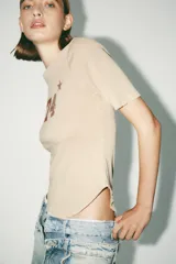 Camiseta color beige de algodón con efecto lavado, corte regular, cuello redondo con tachas metálicas, mangas cortas y estampado frontal con el número 74 y una estrella.