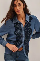 Camisa de denim azul con bordados negros en el frente, de manga larga, cuello clásico y cierre frontal con botones.