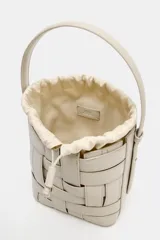 Bolso tipo bucket de diseño trenzado en color marrón oscuro con costuras a la vista en contraste. Cuenta con asa de mano fija, correa bandolera extraíble y compartimento interior de tela en color crudo con cierre mediante fruncido.