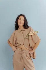 Camisa de lino color tostado, con cuello camisero y cierre frontal de botones. Se caracteriza por sus mangas cortas amplias y voluminosas que aportan un diseño fresco y liviano.