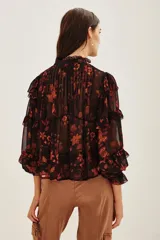 Camisa de viscosa con estampado floral en tonos rojizos y naranjas sobre fondo negro. Presenta volados en los hombros y mangas largas con terminaciones delicadas.