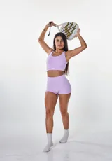 Conjunto deportivo de dos piezas color lila, compuesto por un top deportivo con escote redondo y espalda estilo nadador, y un short de tiro alto ajustado. Ambas prendas presentan el logo de la marca estampado en blanco.