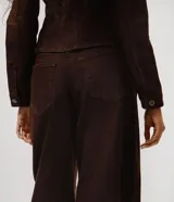 Pantalón de sarga color marrón con corte barrel, cintura media, cierre frontal con botón y cremallera, bolsillos funcionales y recortes estructurales en las piernas.