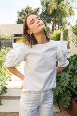 Blusa blanca de algodón con mangas al codo y puños elásticos, con tres volados en las mangas y cierre con botón en la espalda.