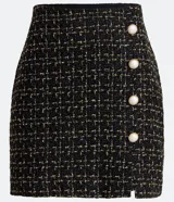 Pollera corta de tweed texturizado en color negro con hilos dorados, presenta botones decorativos con centro blanco y una pequeña abertura frontal en el ruedo.