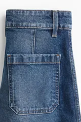Pantalón de jean de corte wide leg, tiro alto con cierre y doble botón frontal. Presenta perneras amplias con pliegues marcados en la parte delantera y trasera, y dobladillo ancho.