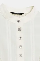 Camisa de rayas finas color beige y blanco, con pechera frontal lisa en tono crudo, cuello mao y volantes aplicados alrededor de la botonadura y los hombros. Cuenta con mangas largas abullonadas y puños rectos a contraste.