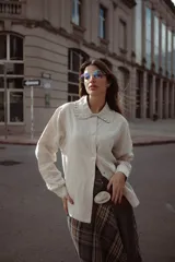 Camisa de lino color beige, con cuello redondo con volados y cierre frontal de botones. Presenta mangas largas con puños abotonados y un corte recto y holgado.