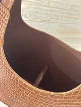 Bolso matero de cuero marrón con textura tipo cocodrilo en la parte superior y cuero liso color beige en la parte inferior. Tiene doble asa de cuero, costuras blancas a la vista y detalles de tachas metálicas. Incluye colgante de cuero y forro interior.