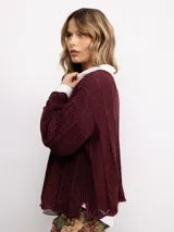 Sweater de punto oversize en color bordó con destellos de lurex. Presenta un diseño acanalado vertical, cuello redondo y mangas largas con terminaciones acampanadas y bordes ondulados.