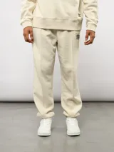 Pantalón jogger de felpa color beige, con corte relajado, costuras frontales verticales y etiqueta con logo de la marca en la parte superior delantera.