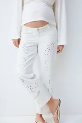 Jean tobillero blanco de corte recto y tiro bajo con detalles de bordado broderie en las piernas. Confeccionado en denim de algodón rígido, cuenta con cinco bolsillos, presillas para cinturón y cierre frontal con botón metálico.