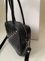 Cartera rígida negra con detalles en rojo y tachas plateadas. Tiene herrajes y cierre metálico plateado marca YKK, asa fina para llevar al hombro o en la mano y correa larga extra regulable. El interior es forrado, tiene un bolsillo con cierre enmarcado en cuero, y un segundo bolsillo plaqué.