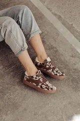 Zapatillas de cuero con estampado que simula pelo de vaca o manchas marrones y blancas. Tienen detalles en cuero marrón liso, cordones marrones y suela de goma color caramelo.
