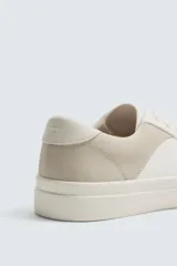 Zapatilla deportiva de corte bajo, color blanco con detalle en tono beige en el talón. Presenta cierre con cordones y suela de goma con volumen.