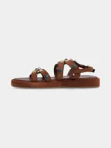 Sandalias planas de cuero gamuza color marrón chocolate, con tiras cruzadas decoradas con apliques metálicos y piedras. Cierre ajustable con hebilla en el tobillo y suela forrada.
