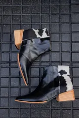 Bota corta de cuero negro con panel trasero de cuero vacuno blanco con manchas negras. Tiene cierre lateral y taco de madera.
