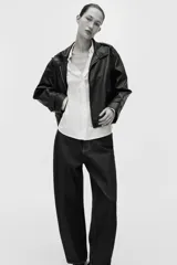 Blusa blanca de tela con brillo, cuello clásico, escote en V con botones frontales, mangas largas con puños sencillos y bajo redondeado.
