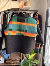 Buzo de cuello alto confeccionado en tejido polar texturizado, con diseño combinado que presenta un sector superior y puños estampados a cuadros en tonos verdes, amarillos, naranjas, celestes y negros, y la parte inferior y mangas en color negro liso.