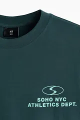 Remera boxy de punto liviano de algodón en color verde oscuro, de hombros caídos, con cuello redondo acanalado y bajo recto. Presenta un diseño estampado en el pecho con texto y logotipo.