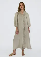 Vestido largo color beige de lino, con cuello mao, mangas 3/4 abullonadas y cierre frontal con botones. Presenta volantes verticales en el frente.