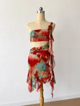Vestido corto de dos piezas (crop top y minifalda) con estampado abstracto en tonos rojos, naranjas, amarillos y azules. El top es tipo bandeau con un tirante que incorpora un volante drapeado que cae sobre el abdomen. La minifalda es ajustada con volante en el ruedo.