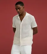 Camisa de manga corta para hombre con cuello solapa y cierre frontal de botones. Presenta un diseño de punto con textura de líneas verticales en color beige y calce regular. Confeccionada en una mezcla de poliéster y algodón sustentable.