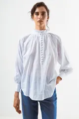 Blusa de algodón blanco con diseño de rayas verticales y textura de pequeños puntos en relieve. Presenta cuello alto con volados, cierre frontal con botones forrados y mangas largas abullonadas con puños ajustados.
