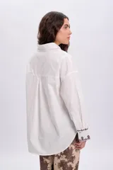 Camisa blanca de corte oversize, con mangas largas y puños anchos decorados con bordados en tono oscuro. Cuenta con cuello camisero clásico y cierre frontal de botones.