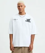 Remera blanca de algodón, corte oversized, con cuello redondo y mangas cortas. Presenta un estampado frontal con un ángel y el texto "PARADISE" en negro, y un pequeño gráfico de estrella/brújula en el lado izquierdo del pecho.