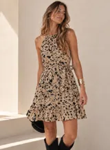 Vestido corto sin mangas con cuello halter, falda con volados y detalle de lazo ajustable en la cintura. Presenta un estampado de manchas abstractas en tonos beige y negro.