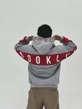 Canguro gris con capucha, de corte holgado, con detalles en color rojo en las mangas y estampado de texto en color rojo en la capucha y el pecho.